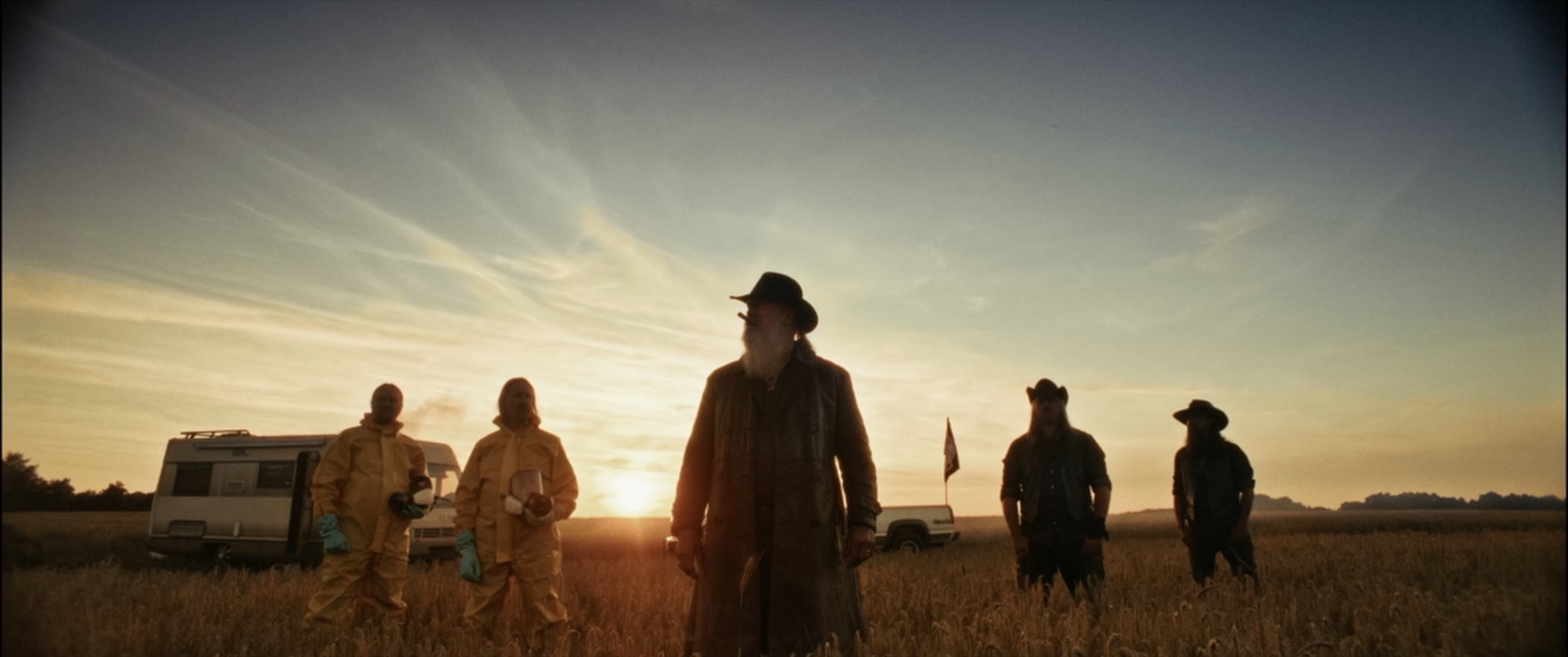 Silhouetted group - two in hazmat suits, a long-coated leader and cowboys - before a low sun in a field - Amon Amarth